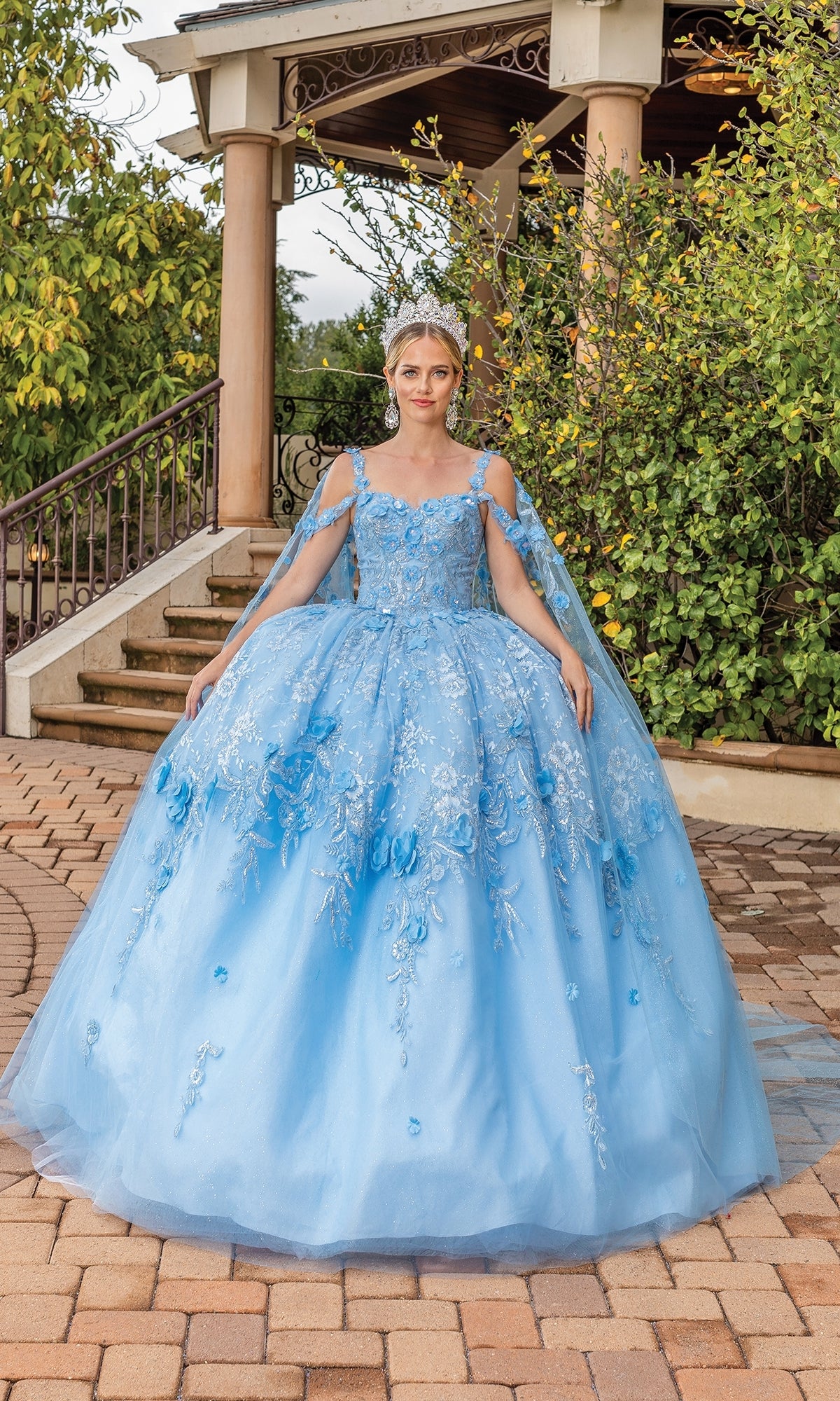 A woman stands outdoors in the Quinceanera Dress 1664 By Dancing Queen, a light blue ball gown with lace embroidered floral details and a silver tiara, posed before garden steps and lush greenery.
